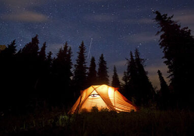 stars above the mountains with tent highlighted by lamp and trees on background